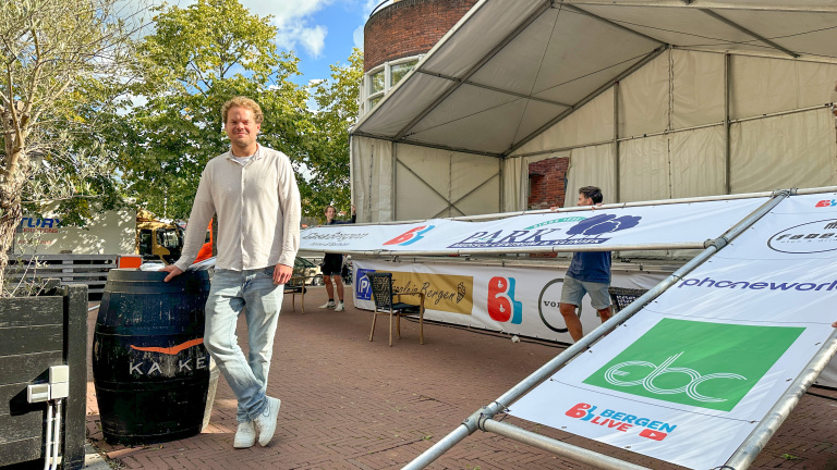 Een man leunt tegen een grote zwarte wijnvat in een buitenruimte met tenten en reclamebanners op de achtergrond. Hij draagt een licht shirt en spijkerbroek. Er zijn bomen en een bakstenen gebouw te zien.