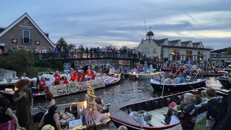 Bootparade op een verlichte gracht met versierde boten en toeschouwers op een brug en langs de kade bij schemering.