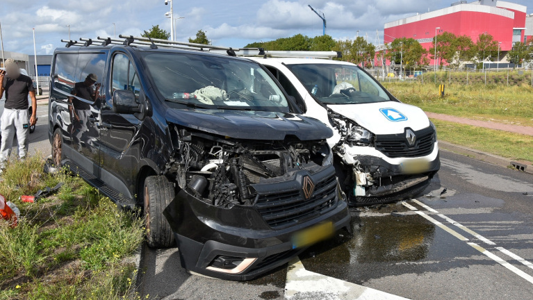 Twee bestelwagens met aanzienlijke schade aan de voorkant na een botsing, geparkeerd aan de kant van de weg met vloeistof op het wegdek. Er staat een man naast het zwarte busje.