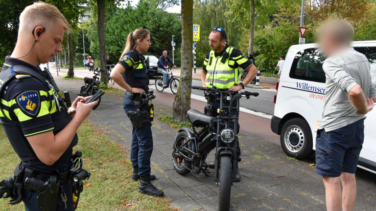 Twee politieagenten in uniform staan op een trottoir en overleggen met een man naast een elektrische fiets; een politiewagen staat geparkeerd op de achtergrond.