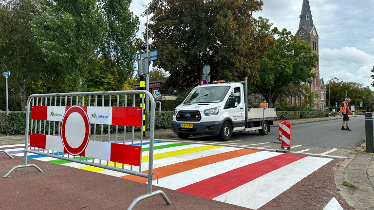 Een regenboogzebrapad in Castricum, afgebakend door een metalen hek met verkeersbord met verbodsbord, een witte bedrijfswagen en een persoon in een oranje veiligheidsvest naast de weg; op de achtergrond een kerk met een toren.