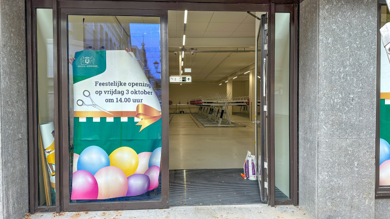 Een openstaande deur naar een lege winkelruimte met een poster op het raam die de feestelijke opening op vrijdag 3 oktober om 14:00 uur aankondigt, versierd met ballonnen en een afbeelding van een schaar en een strik.