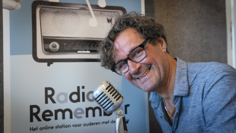 Man met bril en krullend haar lacht naar de camera terwijl hij voor een vintage microfoon zit en naast een poster van 'Radio Remember', een online station voor ouderen.