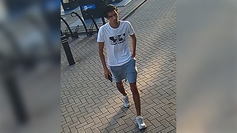 Man met witte T-shirt en korte broek loopt op straat, zijn hand in de zak en telefoon in de andere hand.