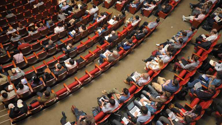 Een zaal vol mensen die in rijen zitten tijdens een conferentie, sommige met laptops en notities.