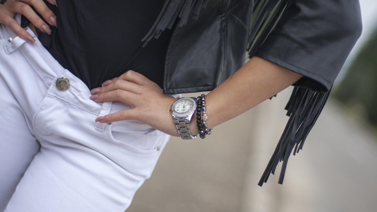 Een persoon draagt witte jeans, een zwart shirt met franjes en een zilverkleurig horloge met armbanden om de pols.