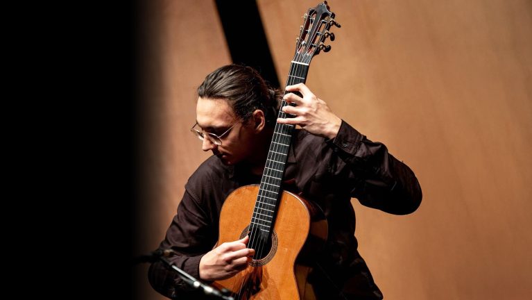 Een persoon speelt een klassieke gitaar met een geconcentreerde houding en bruinachtige achtergrond.