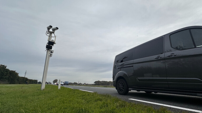 Een donkere bestelwagen rijdt op een landelijke weg, naast een paal met meerdere camera's erop gemonteerd, met een bewolkte lucht op de achtergrond.