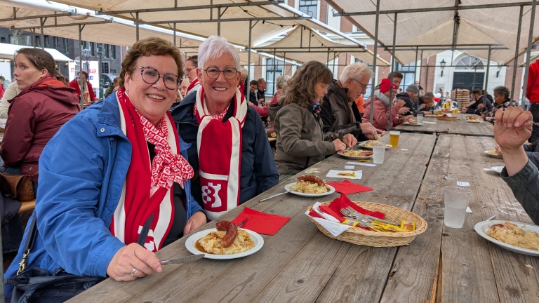 Twee vrouwen dragen rode sjaals met logo's en zitten aan een lange houten tafel, omringd door andere mensen die genieten van een maaltijd met stamppot en worst onder een tentdekking.