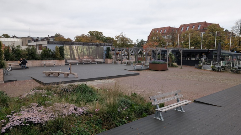 Een plein met houten banken, een speelelement, en beplanting, omgeven door gebouwen en bomen, met een grote afbeelding van een bos aan de muur.