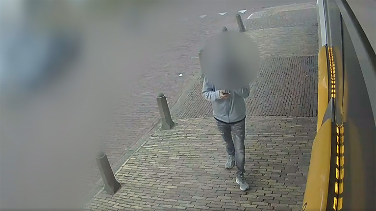 Persoon in een grijs jack en spijkerbroek loopt over een geplaveide straat met een telefoon in de hand, met een gebouw aan de rechterkant en paaltjes langs de weg.