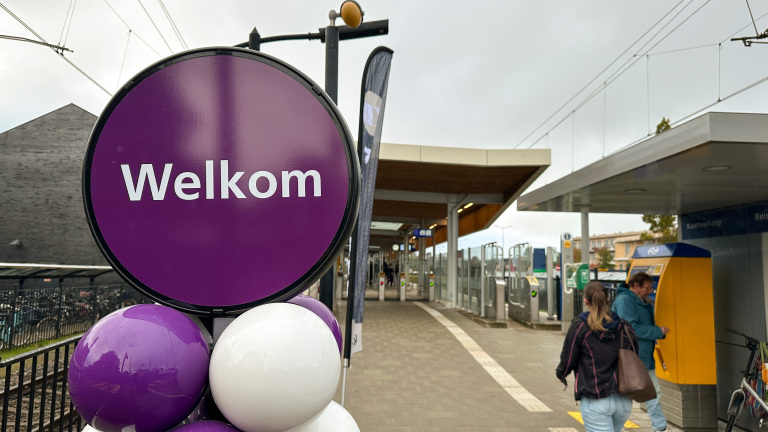 Een paarse welkomstbord staat voor een treinstation met ballonnen en mensen bij een kaartautomaat.