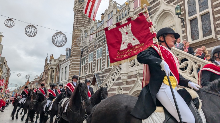 Paardrijders in traditionele kledij met rode sjerpen, een vaandel dragend met referentie aan de "8 October Vereniging Alkmaar Ontzet," marcheren langs een versierde straat met historische gebouwen en toeschouwers.
