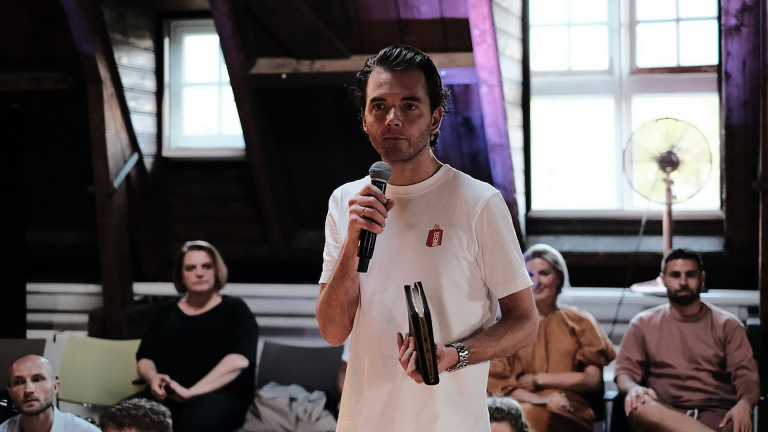 Een man in een witte T-shirt houdt een microfoon vast en spreekt tot een groep mensen die op stoelen zitten in een ruimte met houten wanden en een groot raam.