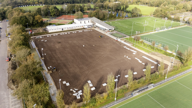 Luchtfoto van een sportcomplex met een groot bruinkleurig veld in aanbouw, omringd door bestaande groene grasvelden en tennisbanen. Er staat een gebouw naast de velden en enkele voertuigen en stenen platen zijn zichtbaar op het bruine veld.