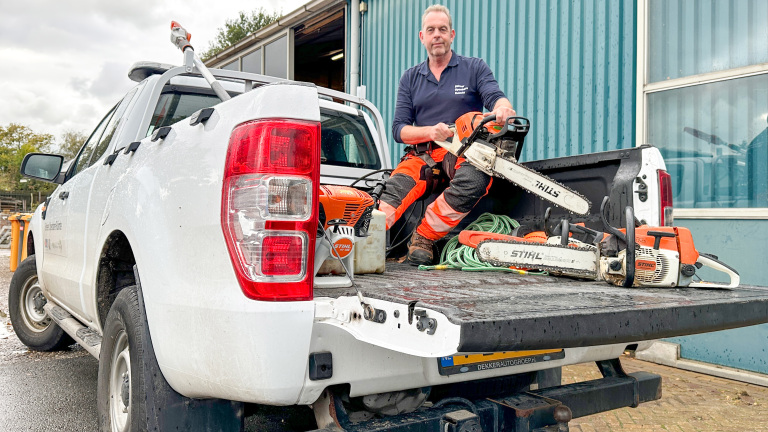 Man in work attire seated on the tailgate of a white pickup truck, holding a chainsaw and surrounded by tools; the truck is parked outside a building with a blue wall.