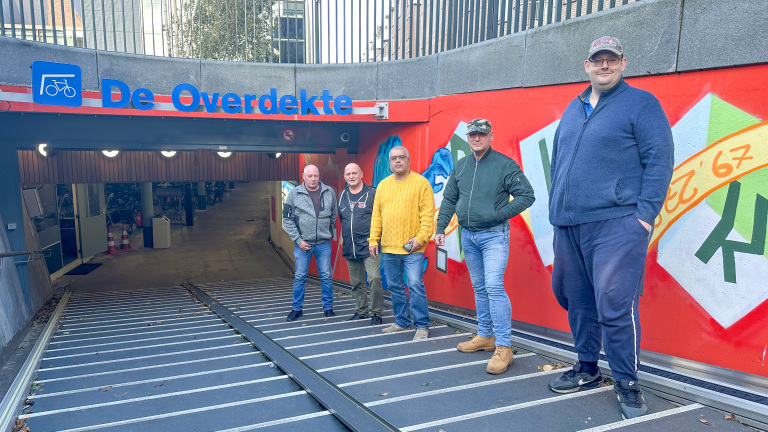 Vijf mannen staan voor de ingang van een fietskelder met de naam 'De Overdekte'. De achtergrond is een helder rode muur met kleurrijke graffiti. De mannen dragen casual kleding en staan aan de zijkant van een betonnen helling die naar de kelder leidt.