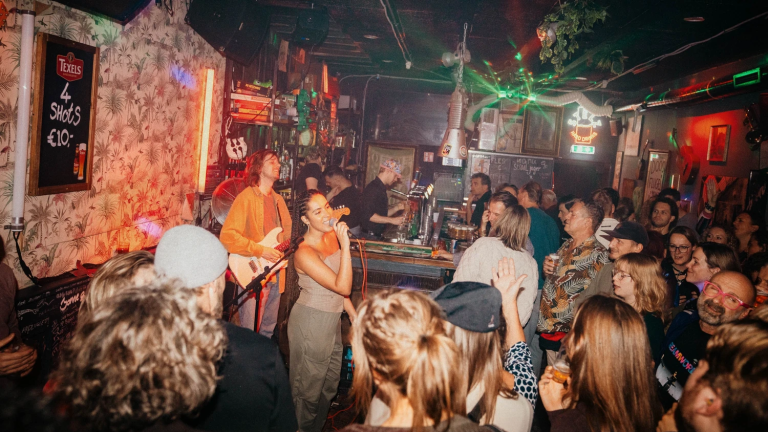 Een zangeres en een gitarist treden op in een drukke bar, omringd door een enthousiast publiek. De sfeer is levendig met kleurrijke verlichting en versieringen aan de muren.
