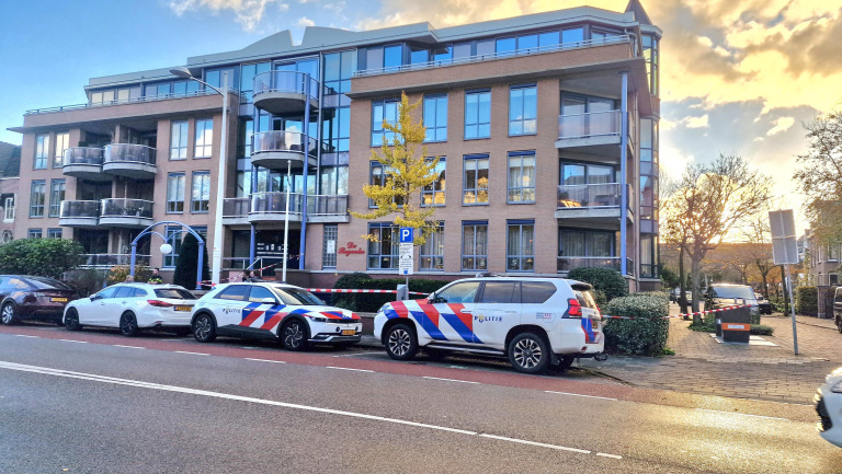 Appartementencomplex met balkons en grote ramen, met twee politieauto's geparkeerd op de stoep ervoor, in de vroege avond met een kleurrijke lucht op de achtergrond.