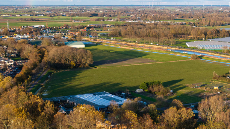 Luchtfoto van een Nederlands landschap met groene velden, een kanaal met een brug, en verspreide gebouwen, omringd door herfstbomen.