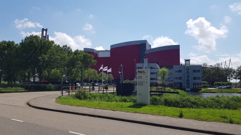 Afvalverwerkingsbedrijf HVC met rode gebouwen, bomen en vlaggen bij de ingang aan een zonnige weg.