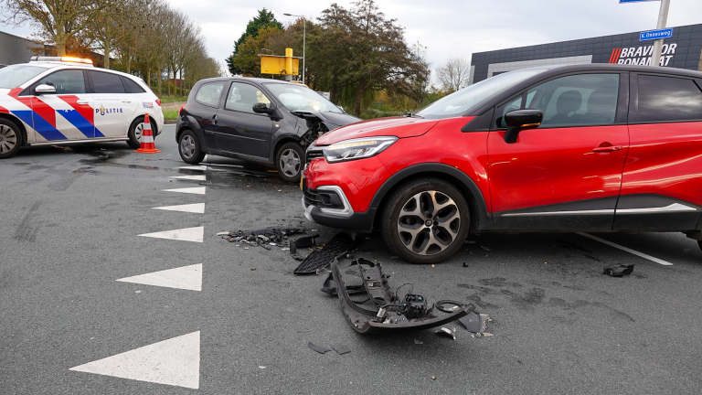 Twee auto's zijn betrokken bij een botsing, met een politieauto die de plaats van het ongeval beveiligt met een verkeerskegel. De voorbumper van de donkere auto is zwaar beschadigd, terwijl brokstukken over de weg verspreid liggen. Op de achtergrond staan bomen en een gebouw met de naam 'Brandoor Donabat'.