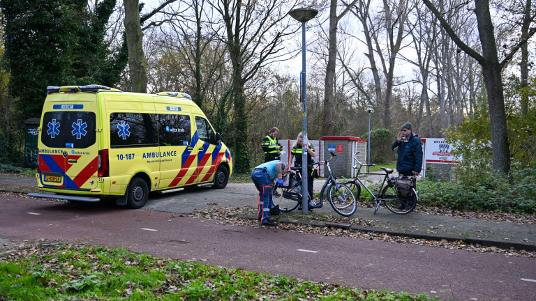 Een gele ambulance staat geparkeerd langs een pad naast een grasveld, terwijl twee politieagenten en een man bij een fiets staan. Een andere man met een fiets staat op de achtergrond. Er zijn bomen en bladeren te zien in de omgeving.