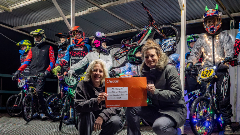 Groep BMX-coureurs met kleurrijke en verlichte kleding en fietsen, geposeerd met twee volwassenen die een cheque van €10.000 vasthouden.