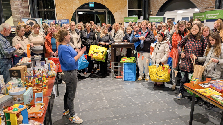Een vrouw in een blauwe trui spreekt voor een publiek in een overdekte ruimte. De aanwezigen luisteren aandachtig en dragen verschillende soorten tassen. Op de tafels vooraan staan kleurrijke speelgoeditems en andere objecten uitgestald.
