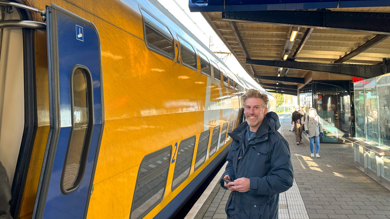 Een man glimlacht bij een gele NS dubbeldekker trein op een perron.