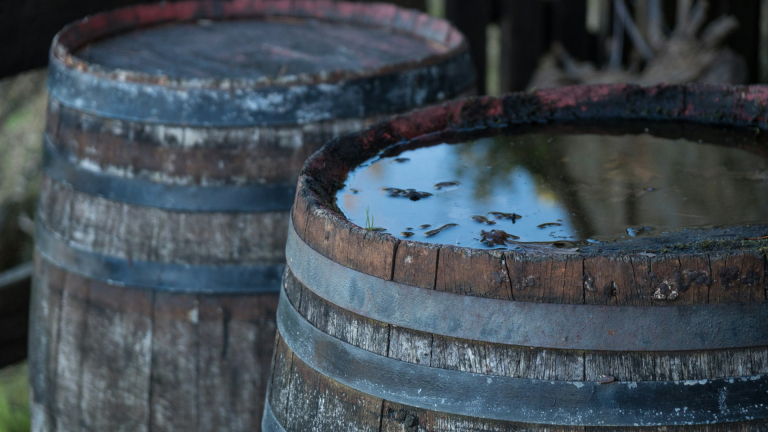 Twee oude houten vaten waarvan één gevuld is met stilstaand water op de bovenkant, geplaatst in een rustieke omgeving.