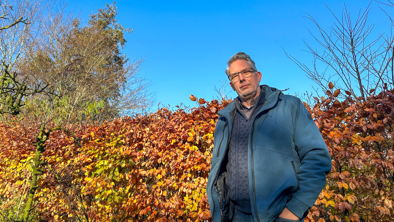 Man met bril en blauw jack staat voor een haag met herfstbladeren onder een blauwe hemel.