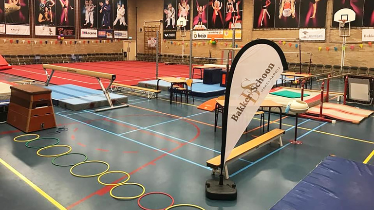 Een gymzaal met diverse gymnastiektoestellen zoals een evenwichtsbalk, springkast, en ringen op de vloer, met op de achtergrond reclameposters aan de muur.