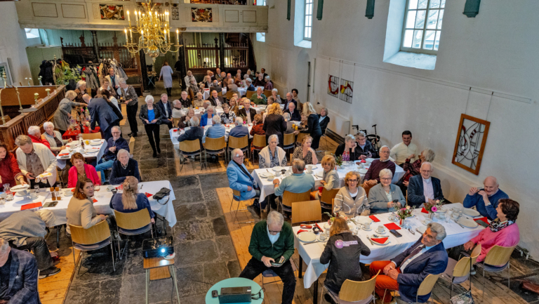 Groep mensen zit aan tafels in een kerkzaal tijdens een bijeenkomst, met witte tafelkleden en bloemen op de tafels en een kroonluchter bovenaan.