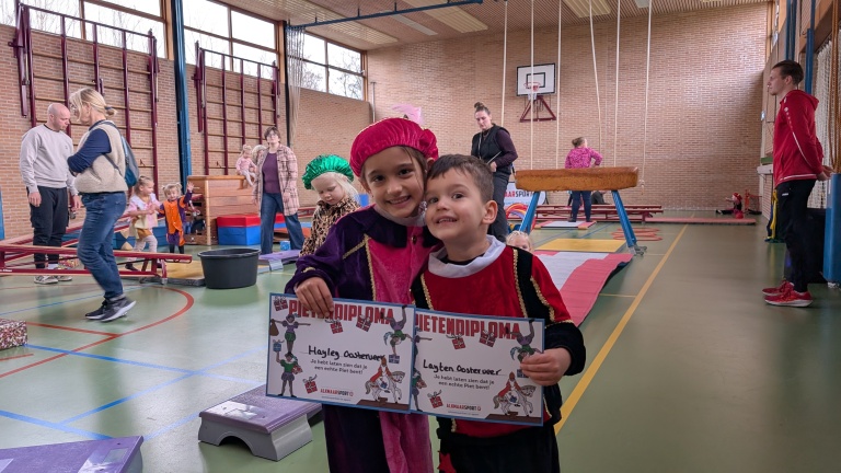 Twee kinderen in kleurrijke kostuums houden hun Pieten diploma's vast in een sportzaal gevuld met mensen en sportapparatuur.
