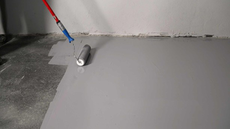 Een verfroller met een rode en blauwe steel wordt gebruikt om een grijze vloer te verven in een binnenruimte.