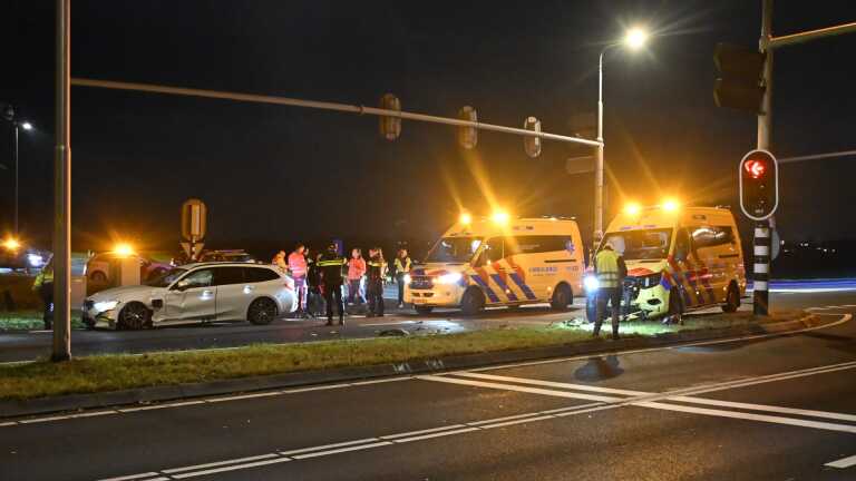 Een verkeersongeval in de nacht met een beschadigde witte auto op de weg en meerdere hulpverleners en ambulances aanwezig, gefotografeerd onder straatverlichting.