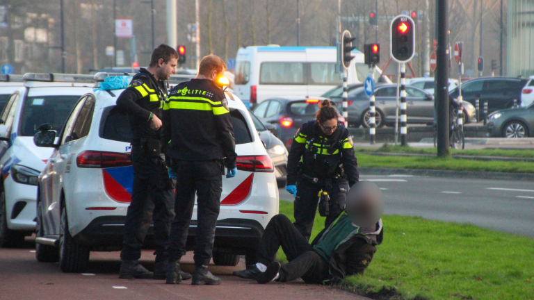 Drie politieagenten in uniform assisteren een liggende man op de stoep naast een politieauto, met verkeer en stoplichten zichtbaar op de achtergrond.