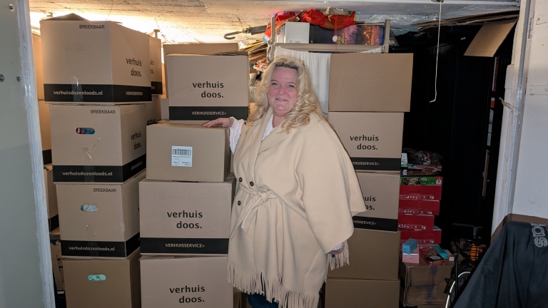 Vrouw met blond haar staat in een ruimte volgestapeld met verhuisdozen, gekleed in een beige jas.