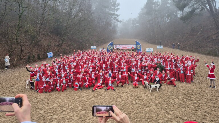 Een grote groep mensen in kerstmanpakken poseert op een zandpad, met bos en een bord met de tekst "Kerstmannenrun".