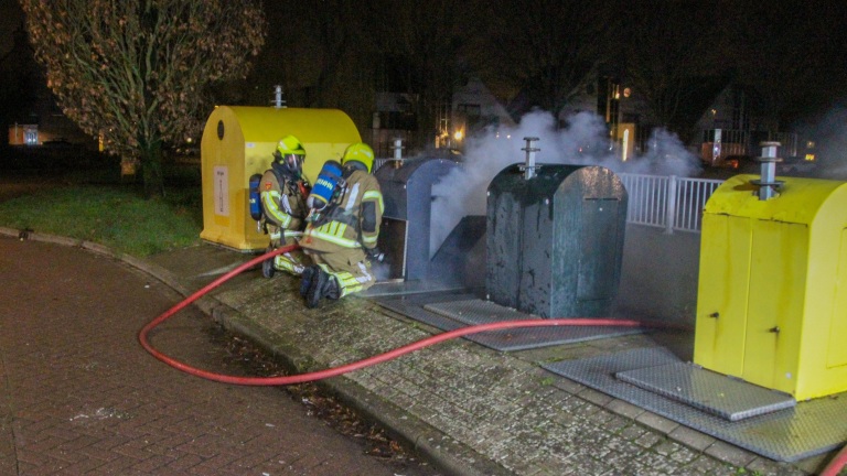 Brandweerlieden blussen 's nachts een brand in een ondergrondse afvalcontainer, met rook die uit de container komt.