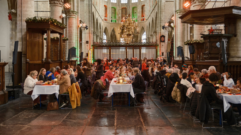 Mensen zitten aan lange tafels in een kerkelijke omgeving, bezig met een maaltijd tijdens een evenement.