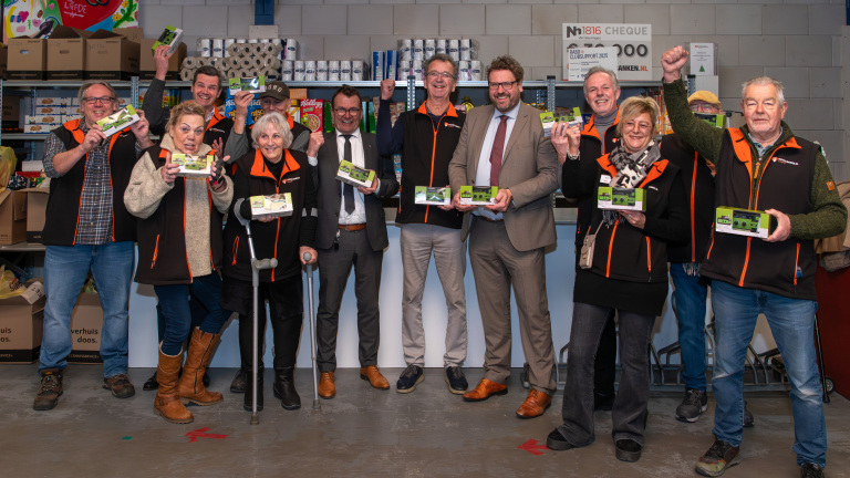 Een groep mensen poseert enthousiast met producten in een magazijn met planken vol levensmiddelen.