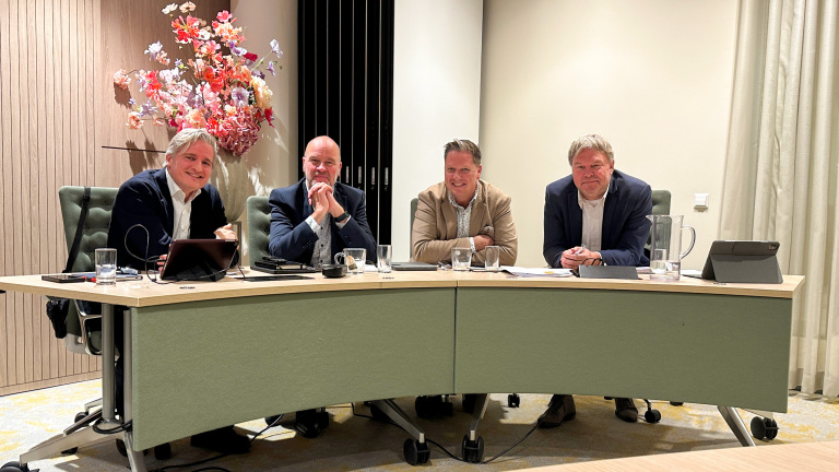 Vier mannen zitten aan een tafel in een vergaderruimte, met bloemen achter hen en elektronische apparaten op tafel.