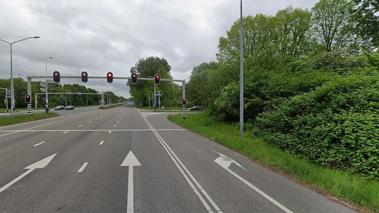 Verkeerslicht op rood boven een brede, lege weg met rijbanen gescheiden door groene bermen en omgeven door bomen.