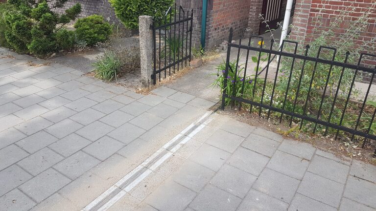 Een trottoir met grijze tegels, een laag hekwerk, en een kleine tuin met planten voor een bakstenen huis.