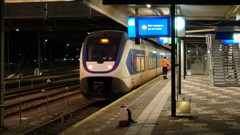 Een passagierstrein bij een perron met een bord "Niet instappen"; een werknemer in oranje jas loopt langs het perron.