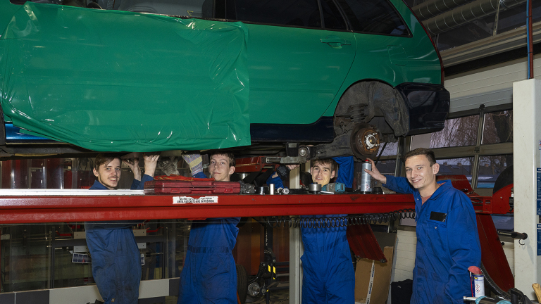 Vier mannen in blauwe overalls werken onder een auto die op een hefbrug staat in een garage.
