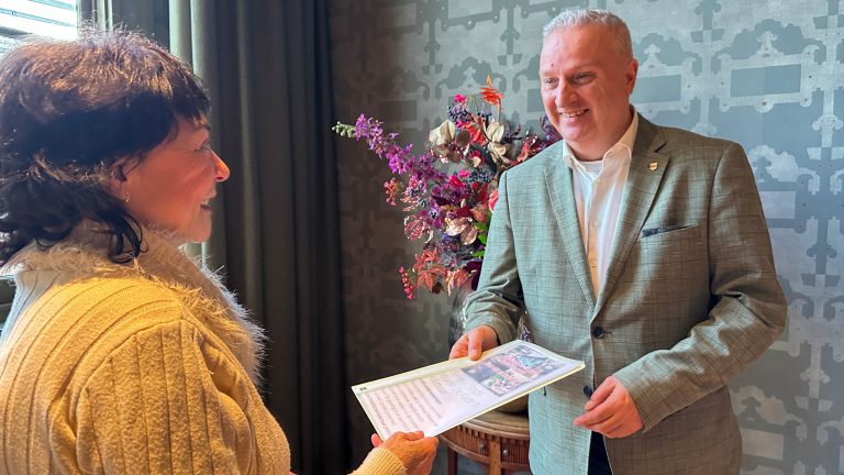 Een lachende man in een grijze blazer overhandigt een certificaat aan een vrouw in een beige trui, met bloemen op de achtergrond.