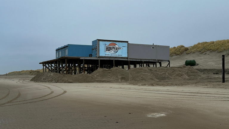 Strandpaviljoen op palen met een bord "Hookipa Beach" tegen een achtergrond van duinen.